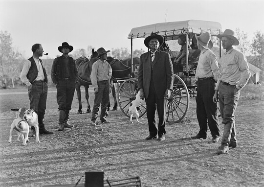 A black-and-white image of six men dressed in western attire and several dogs standing in front of a carriage.
