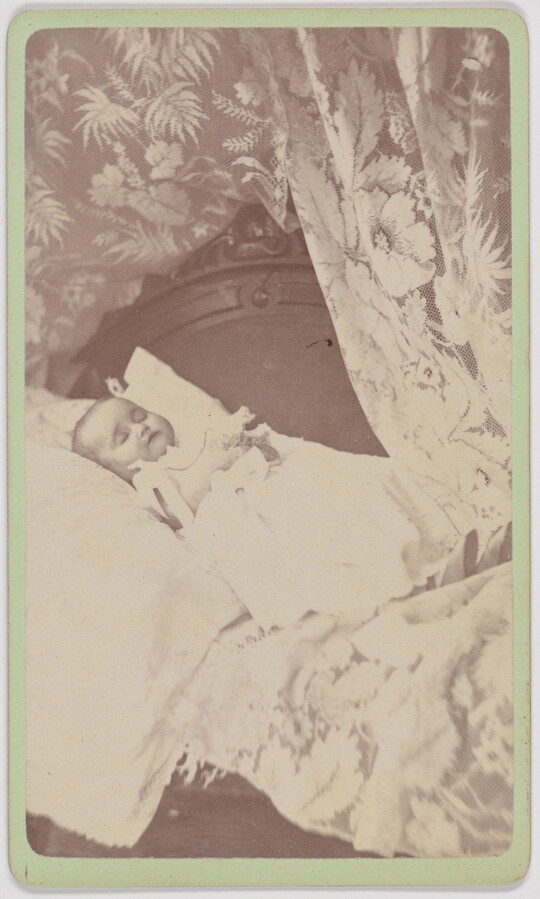 A black-and-white photograph of a White baby wearing a white gown and positioned on a pillow framed by a floral curtain.