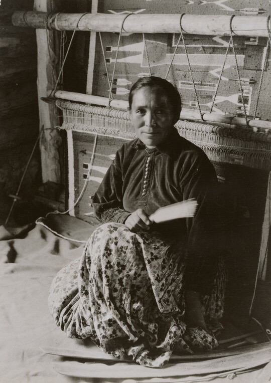 A black-and-white photographic portrait of a Native American woman sitting on the floor in front of a large loom.