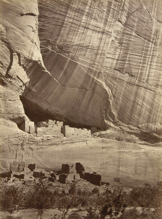 A sepia-toned photograph of cliff dwellings at the base of a large, sheer cliff.