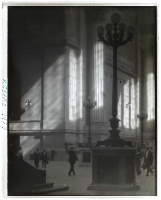 A black-and-white photograph of people walking through a sunlit plaza with towering light posts and columns.
