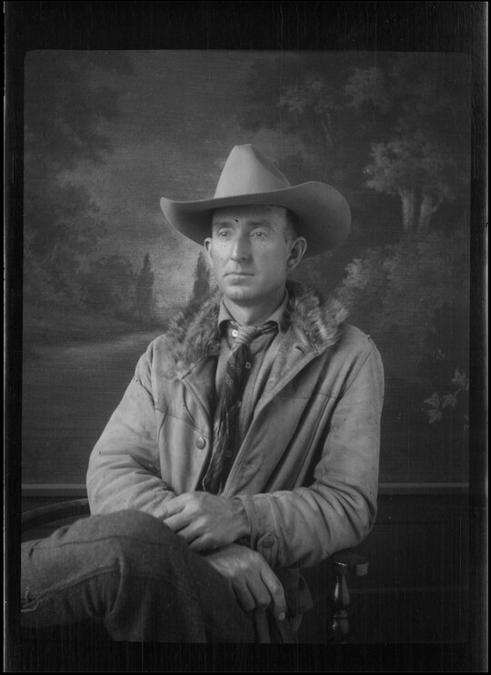 A black-and-white image of a seated White man wearing a cowboy hat, coat, and tie in front of a painted landscape.