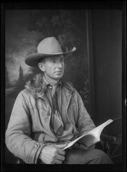 A black-and-white image of a seated White man wearing a cowboy hat, coat, and tie, holding an open book on his lap.