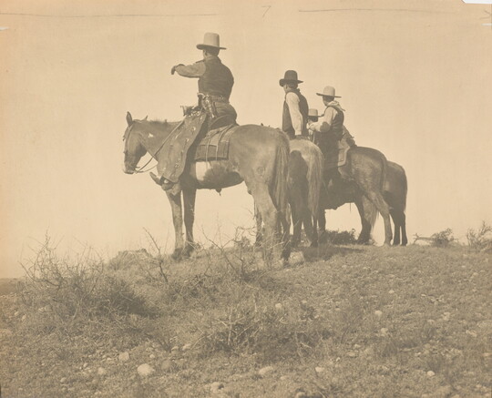 A sepia-toned photograph of three cowboys, one gesturing to the distance, on horseback in a field.