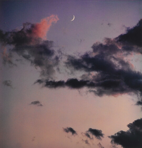 A color photograph of a crescent moon at dusk above thin, dark clouds.