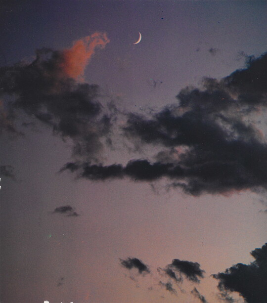 A color photograph of a crescent moon at dusk above thin, dark clouds.