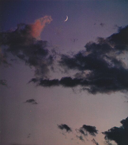 A color photograph of a crescent moon at dusk above thin, dark clouds.