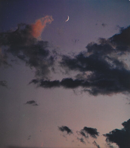 A color photograph of a crescent moon at dusk above thin, dark clouds.