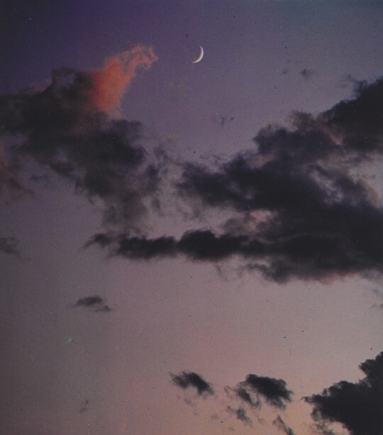 A color photograph of a crescent moon at dusk above thin, dark clouds.