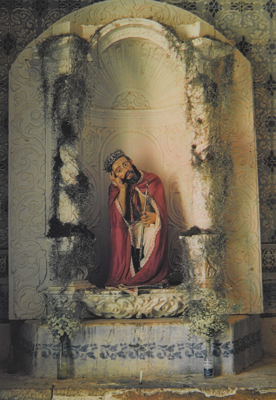 A color photograph of a painted sculpture of a seated Jesus wearing a crown, red robe, holding a cane, head resting on his hand in an arched niche.