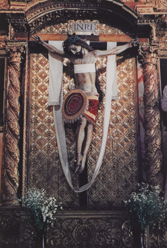A color photograph of a painted sculpture of a crucified Jesus wearing a crown of thorns, a white cloth draped over the cross, hanging on an ornate gold wall.