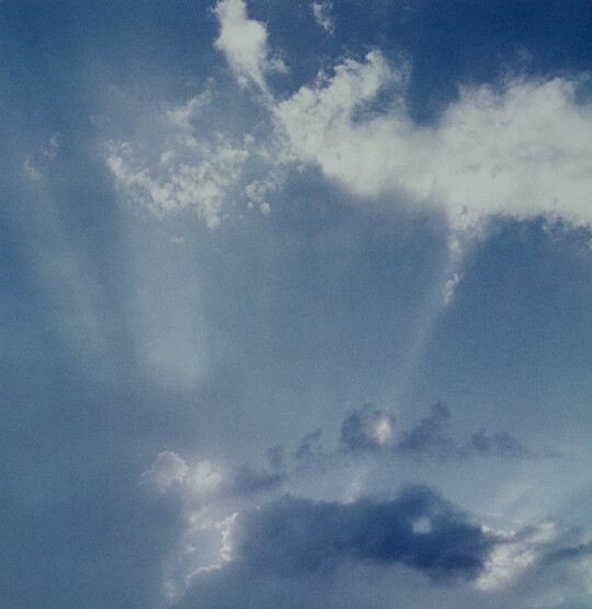A color photograph of white clouds in a blue sky and sun rays that create white streaks.