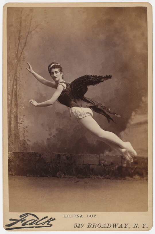A sepia-toned studio photograph of a White woman, black wings on her back, arms extended, suspended in the air as if flying.