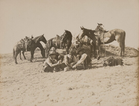 A sepia-toned photograph of a group of men sitting outside on the ground playing cards as their saddled horses stand behind them.