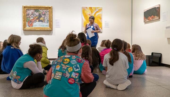 A group of Girl Scouts sit on the floor in a gallery and look at an educator. 