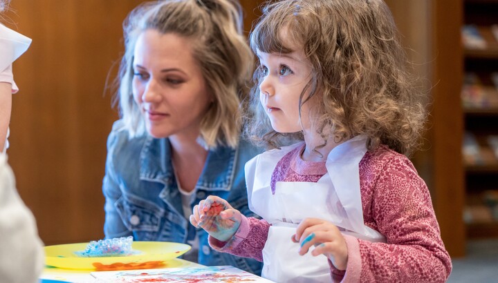 A toddler with paint-covered hands and an adult make art at a table.
