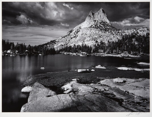 A black-and-white photograph of a rocky mountain peak from across a still lake surrounded by trees.