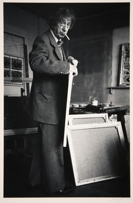 A black-and-white photograph of an older White man with a cigarette in his mouth holding a canvas in an artist's studio.