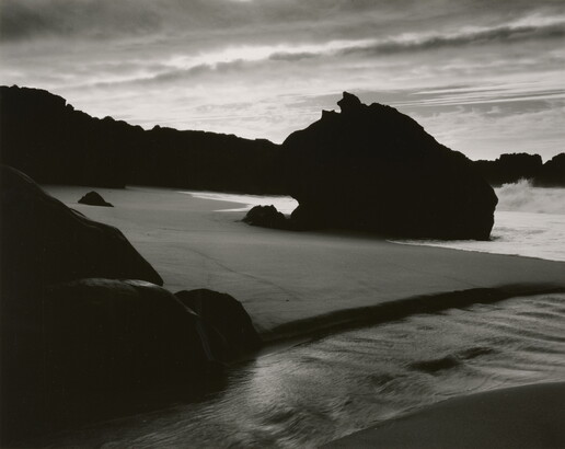 A black-and-white photograph of the sand and sea next to a shore covered with large rocks.