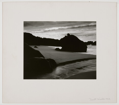 A black-and-white photograph of the sand and sea next to a shore covered with large rocks.
