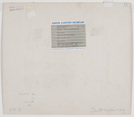 A black-and-white photograph of the sand and sea next to a shore covered with large rocks.