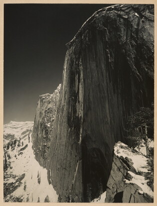 A black-and-white photograph of a steep cliff face and the snow-covered mountains around it.