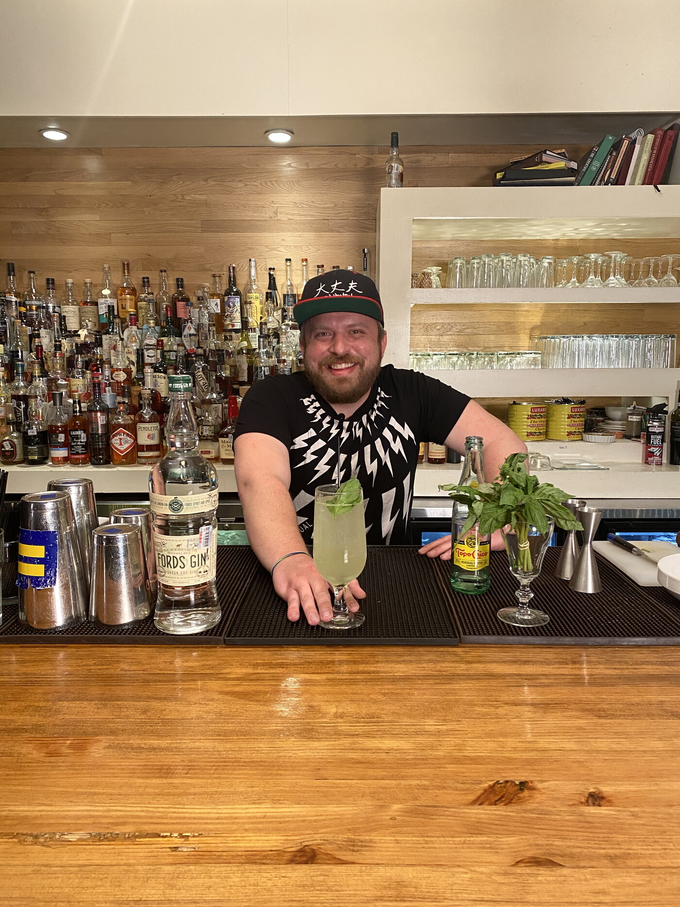 A smiling person pushes a mixed drink in a pilsner forward on a bar.