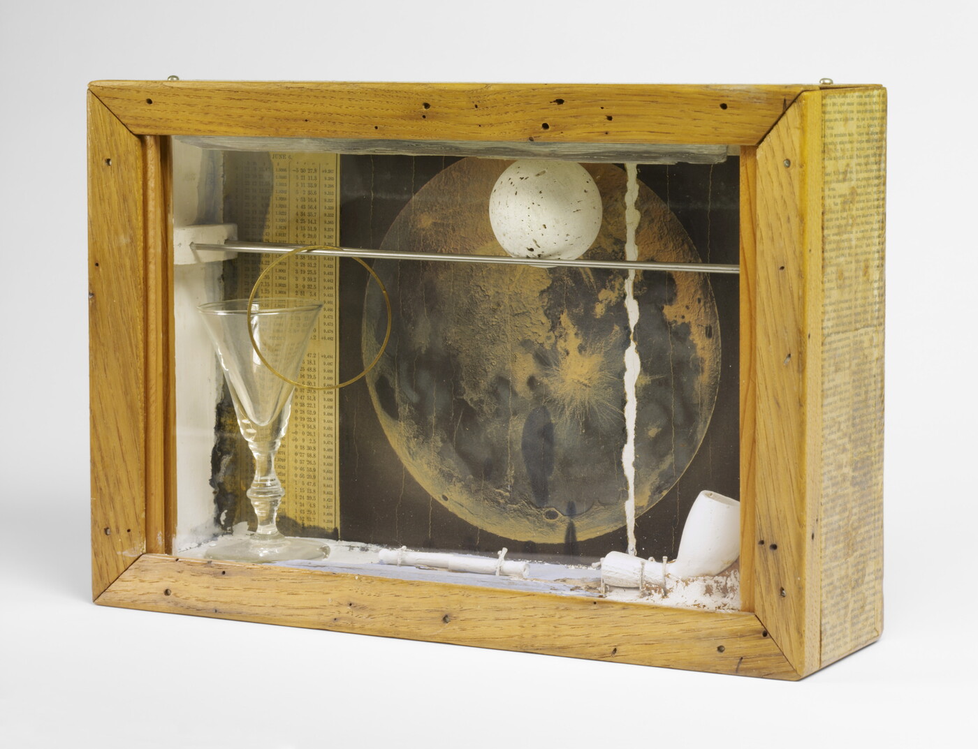 A wood shadowbox containing a pipe, a white ball with gray speckles, a picture of the moon, and a glass goblet.