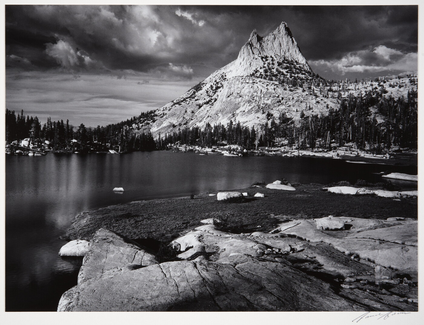 A black-and-white photograph of a rocky mountain peak from across a still lake surrounded by trees.