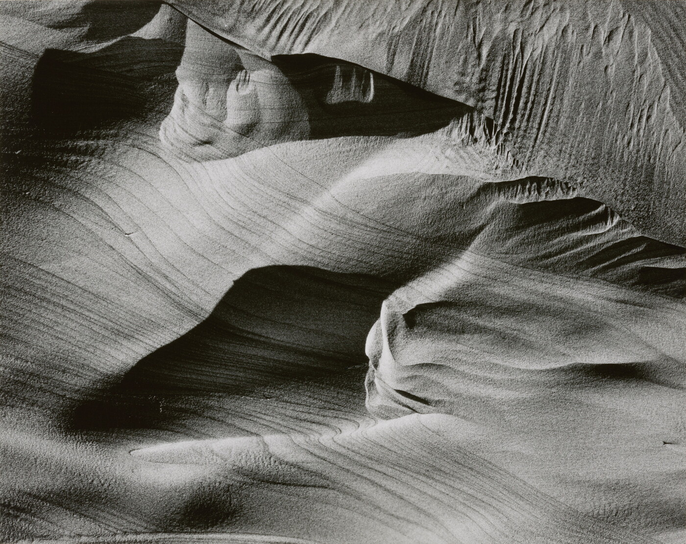 An abstract black-and-white photograph of sand dunes with horizontal and vertical striations that make it seem to be in motion.
