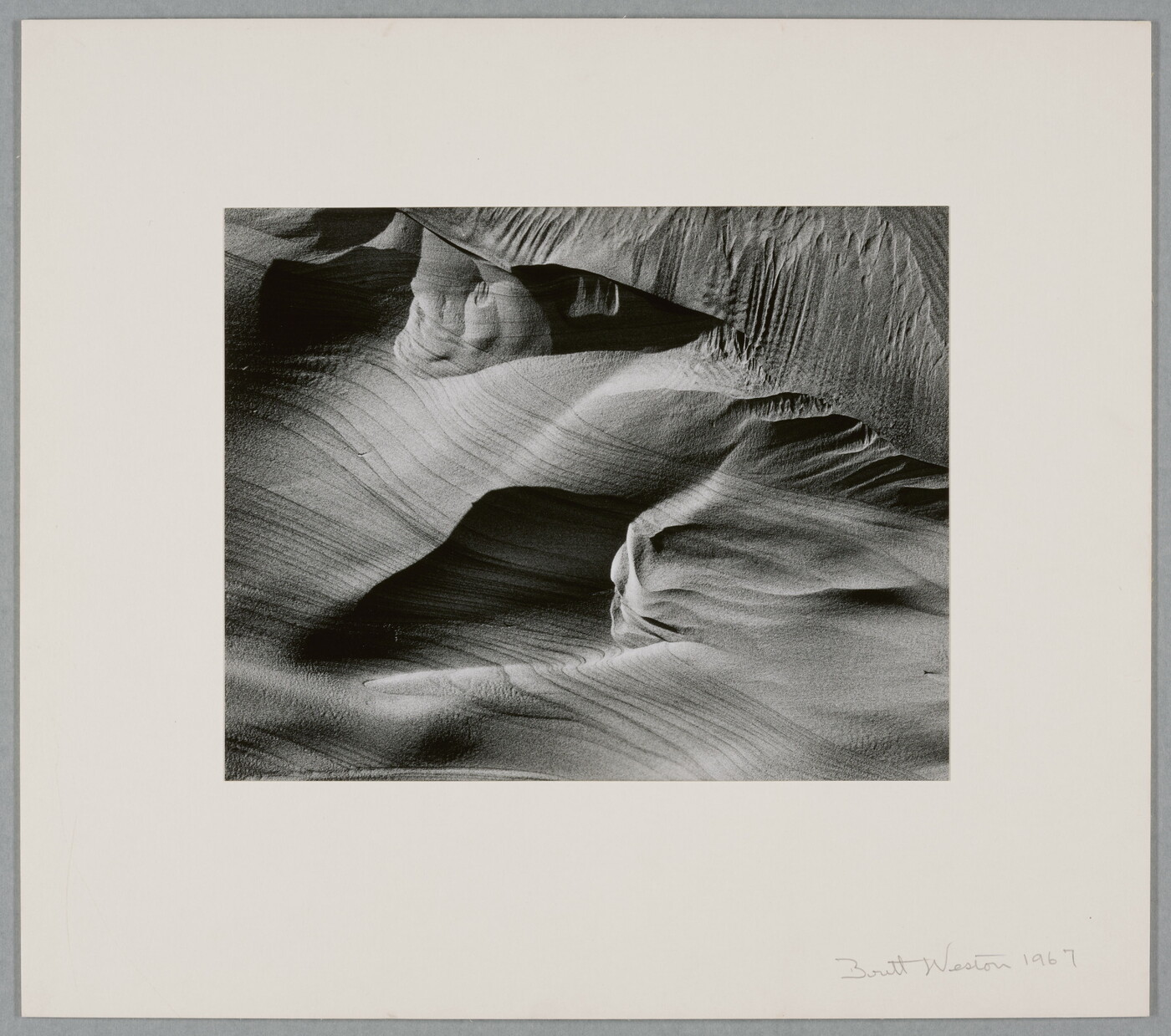 An abstract black-and-white photograph of sand dunes with horizontal and vertical striations that make it seem to be in motion.