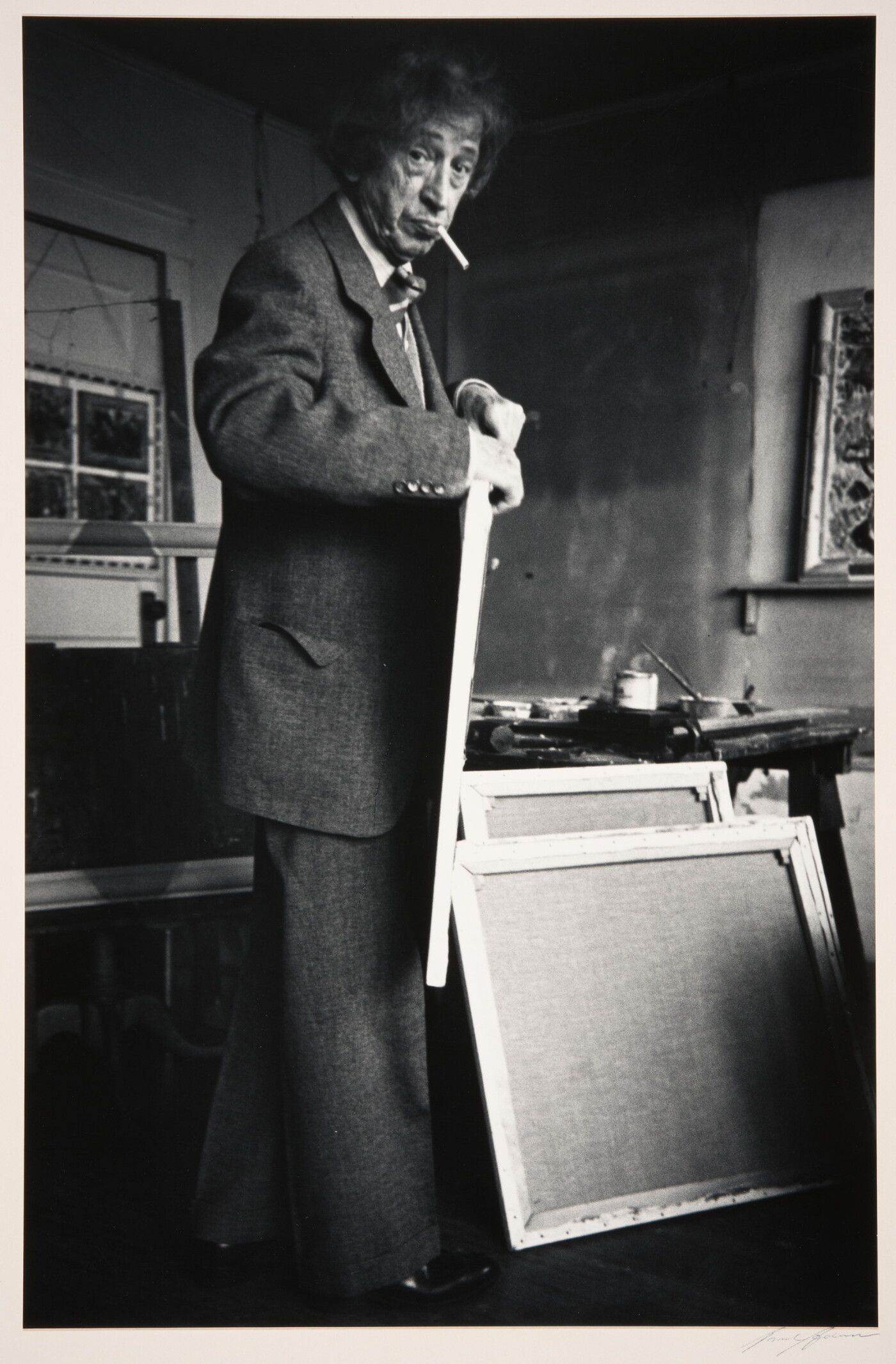 A black-and-white photograph of an older White man with a cigarette in his mouth holding a canvas in an artist's studio.