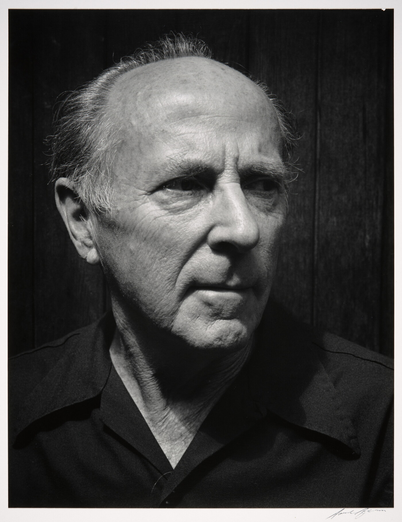 A black-and-white photograph of the head and shoulders of an older balding White man wearing a dark button-down shirt, head turned slightly.