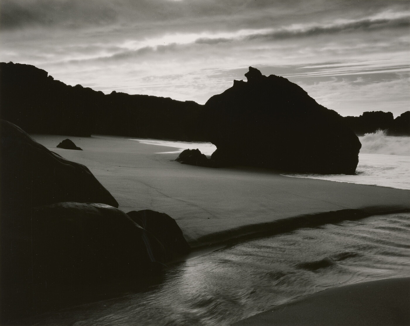 A black-and-white photograph of the sand and sea next to a shore covered with large rocks.