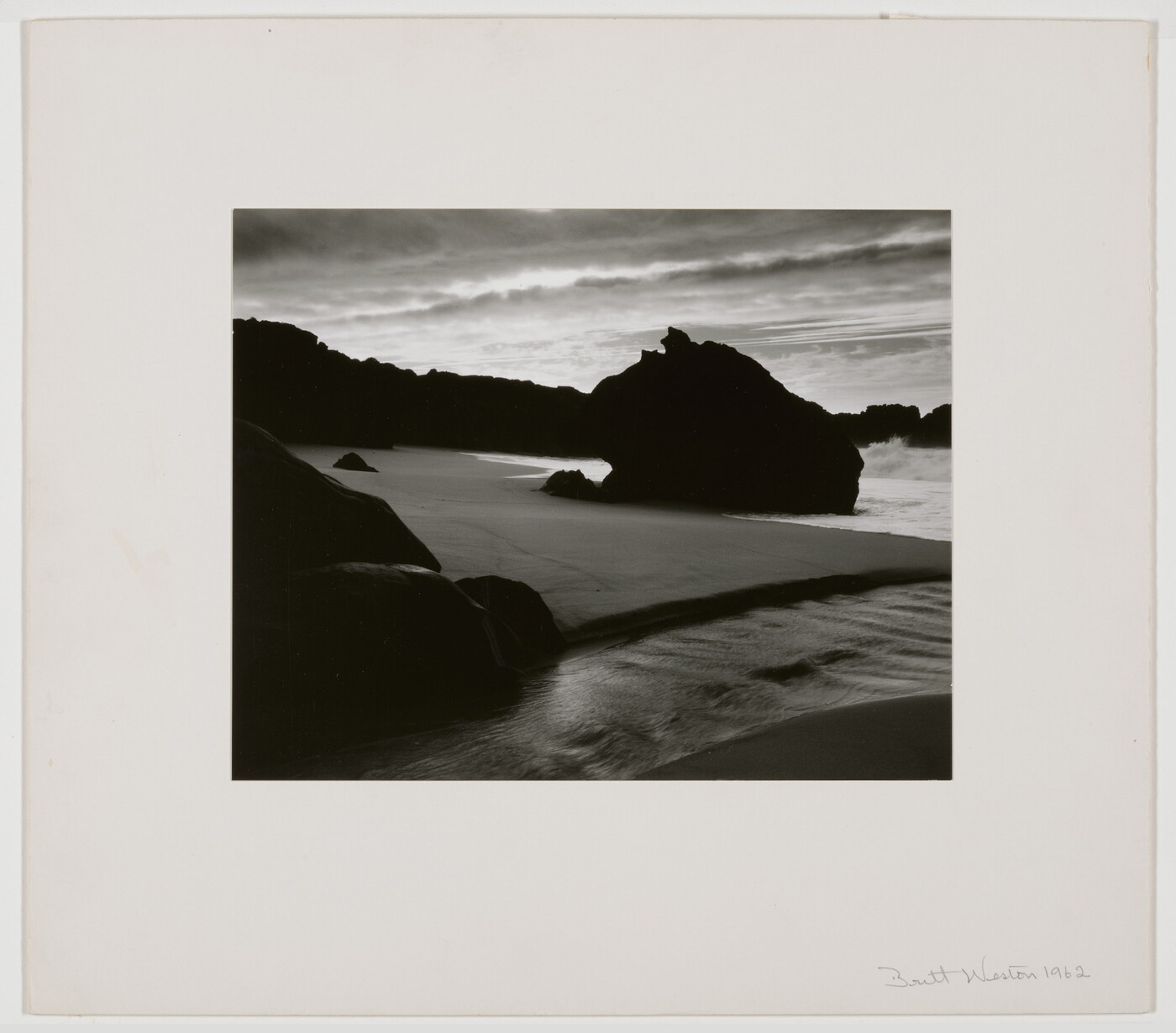 A black-and-white photograph of the sand and sea next to a shore covered with large rocks.