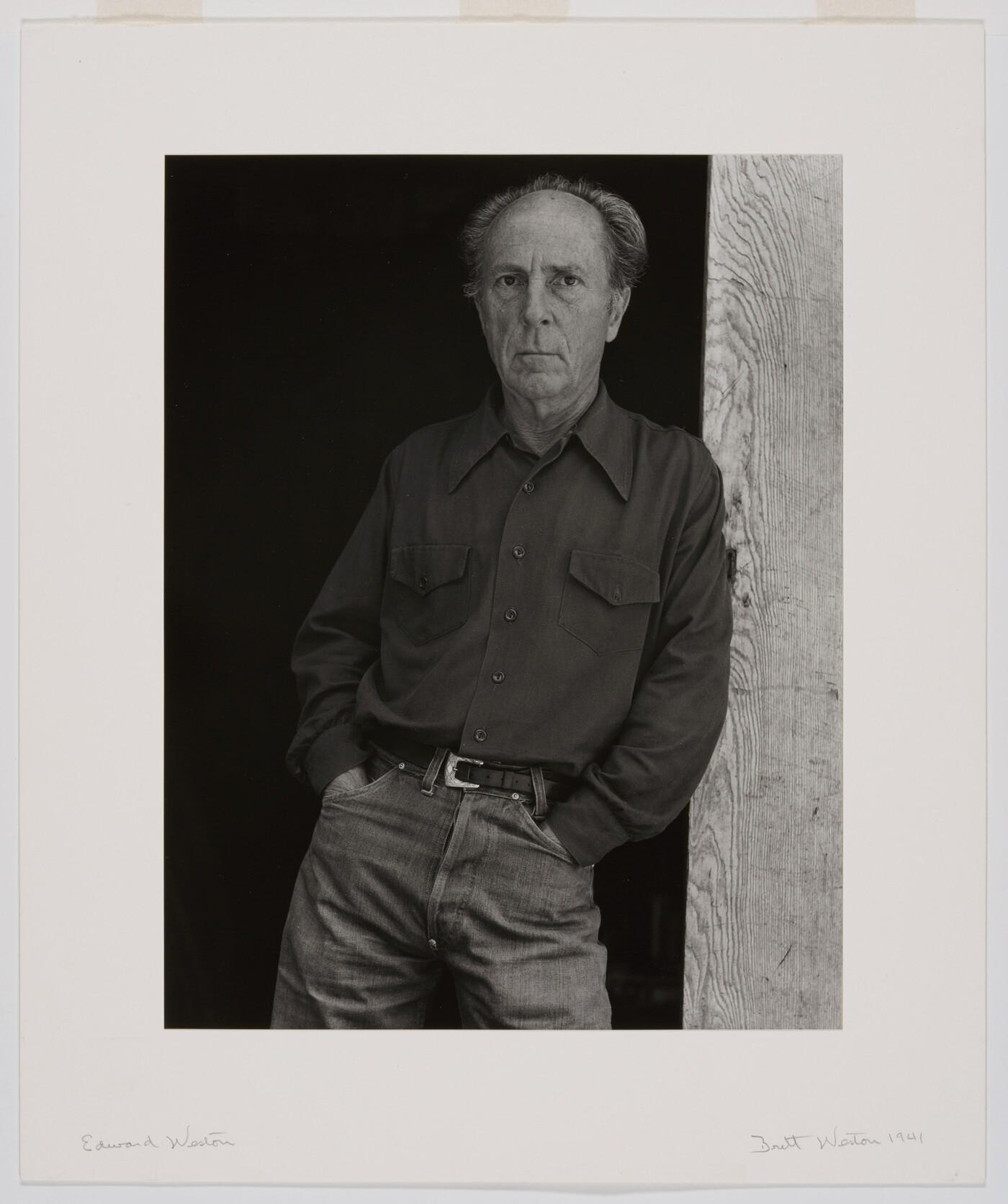 A black-and-white portrait of an older, balding White man wearing jeans and a long-sleeve button-down shirt leaning against a wooden beam.
