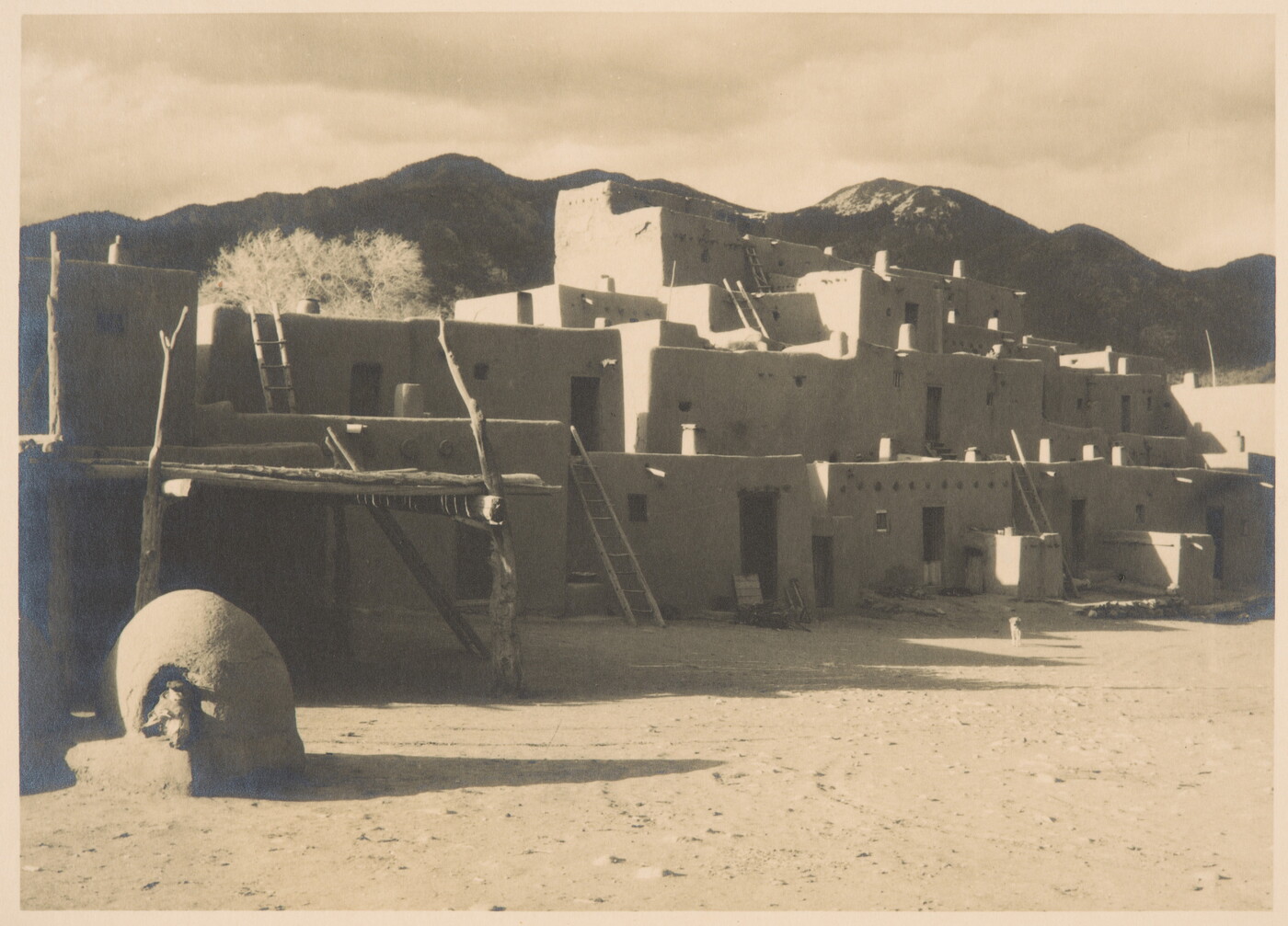 A black-and-white photo of a multi-level adobe pueblo with ladders leading from story to story and dark mountains in the background.