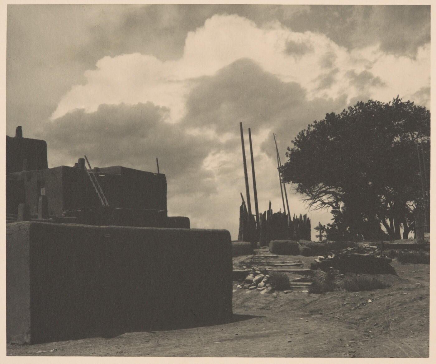 A black-and-white photograph of multi-level adobe pueblo with tall, wood poles standing upright in the distance under a cloudy sky.