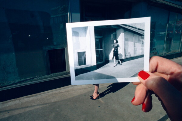 A color photograph of a White hand with red fingernails holding a black-and-white photo of a Woman walking in front of empty storefronts up in front of empty storefronts.
