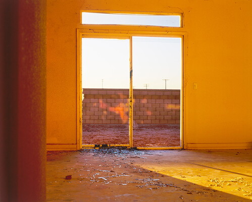 A color photograph looking through the sliding doors, one with a broken window, of a yellow room out onto a brick wall.