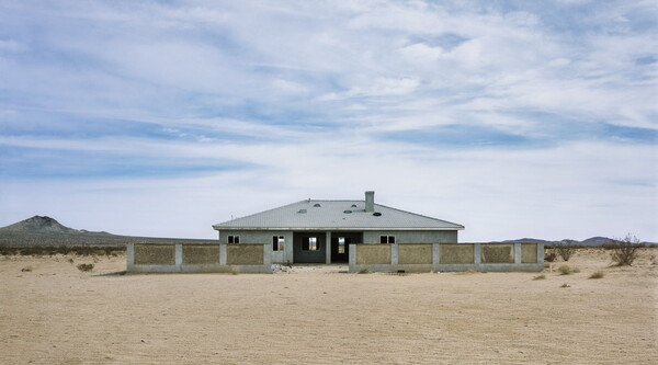 A color photograph of a one-story building that is under construction behind a brick wall in a desert landscape.