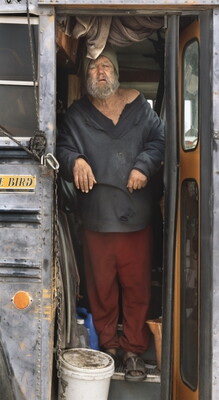 A vertically-oriented color photograph of an older White man with gray hair and beard wearing disheveled clothes standing the the doorway of a school bus.