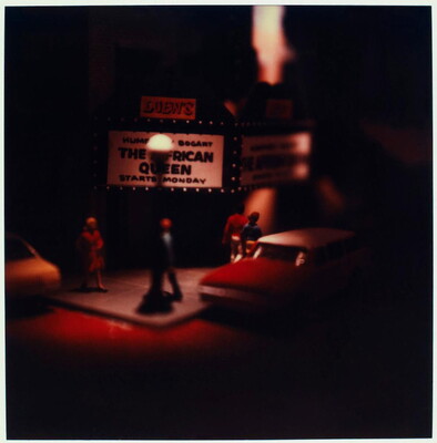 A very soft-focus color photograph of a model of a street corner movie theater with cars parked nearby and people on the sidewalk.