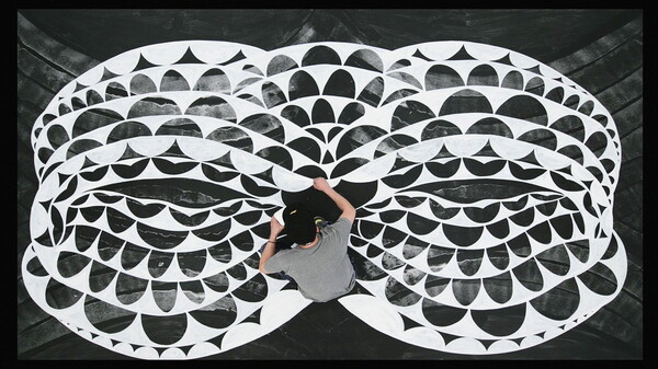 A color photograph of a White man, as viewed from above, sitting on a floor painting a large black-and-white design.