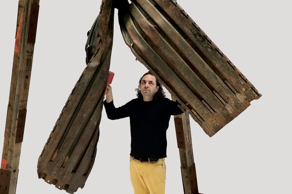 A medium-skinned man with dark curly hair stands under two large pieces of metal with vertical slits in them/