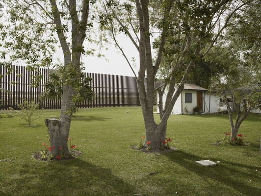 A color photograph of a small white house, trees in a well-manicured yard, and a tall fence at the back of the yard.
