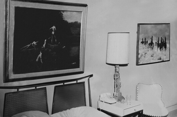 A black-and-white photograph of a hotel room showing a painting above the bed and another painting hanging above a side table.