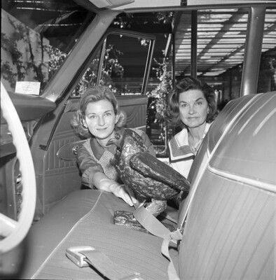 A black-and-white photograph of two White women kneeling next to the open passenger door of a car placing a seatbelt on a sculpture of an owl.