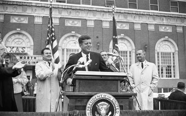 A black-and-white photograph of President John F. Kennedy speaking at a podium in front of a brick building.