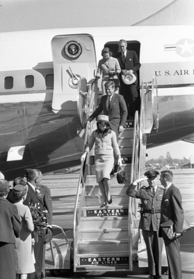 A black-and-white photograph of John F. Kennedy and Jacqueline Kennedy descending the stairs from an airplane as people on the tarmac wait to greet them.